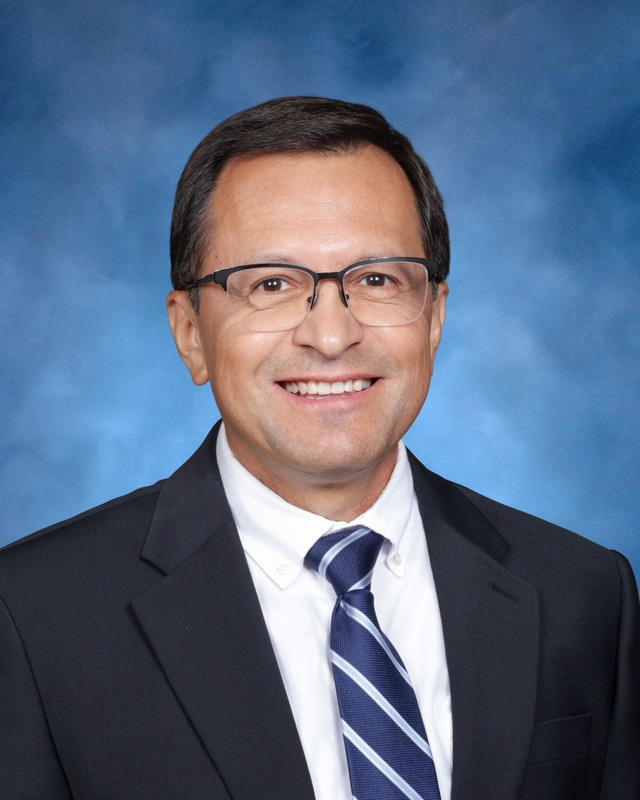 A man wearing glasses, a dark suit, white shirt, and striped tie smiles in front of a blue background.