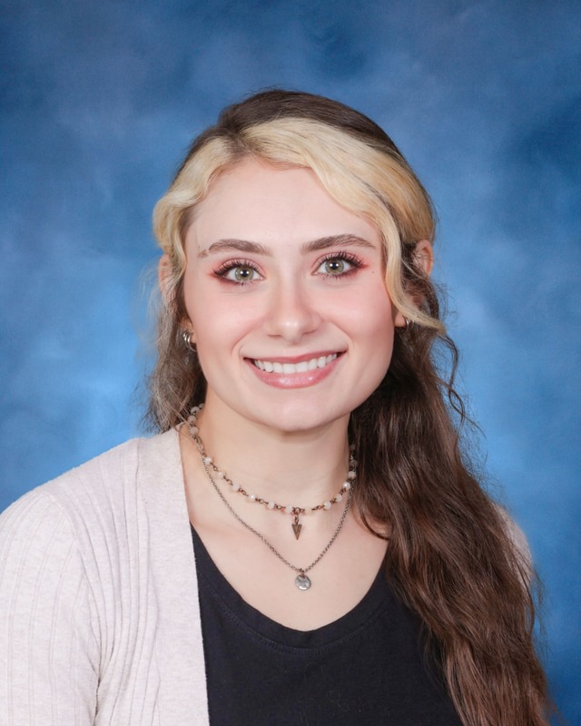 A young woman with long, wavy brown and blonde hair smiles at the camera in front of a blue studio background.