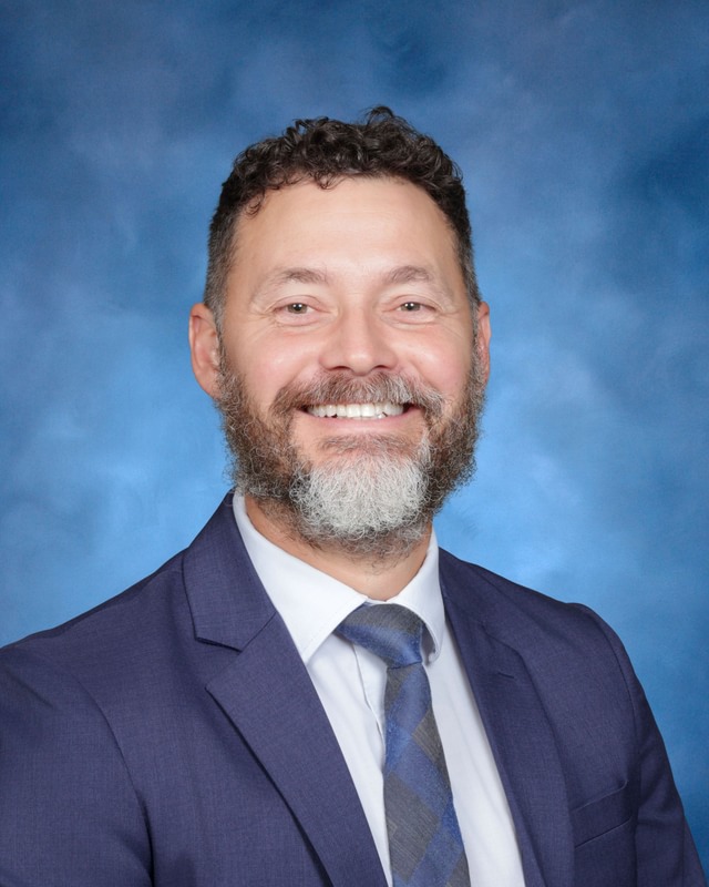 A man with curly hair and a beard smiles, wearing a blue suit, white shirt, and striped tie, against a blue studio background.