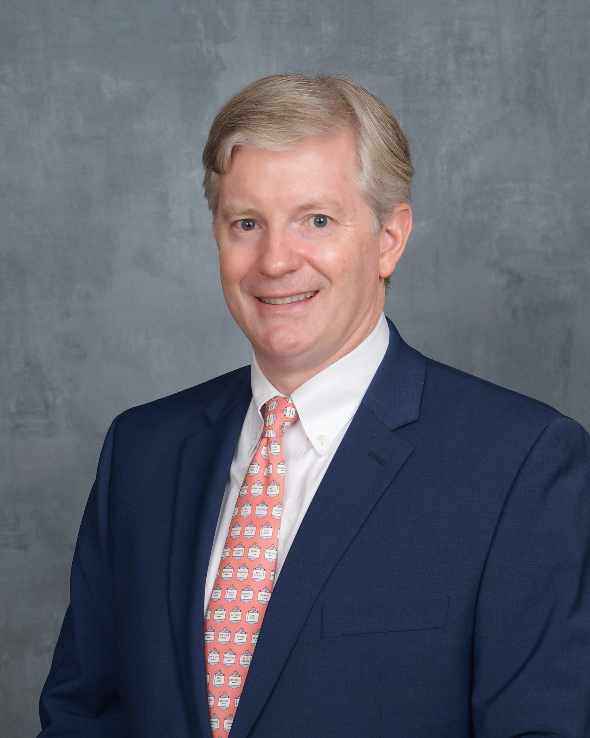 Man in a blue suit and patterned pink tie smiling at the camera against a gray background.