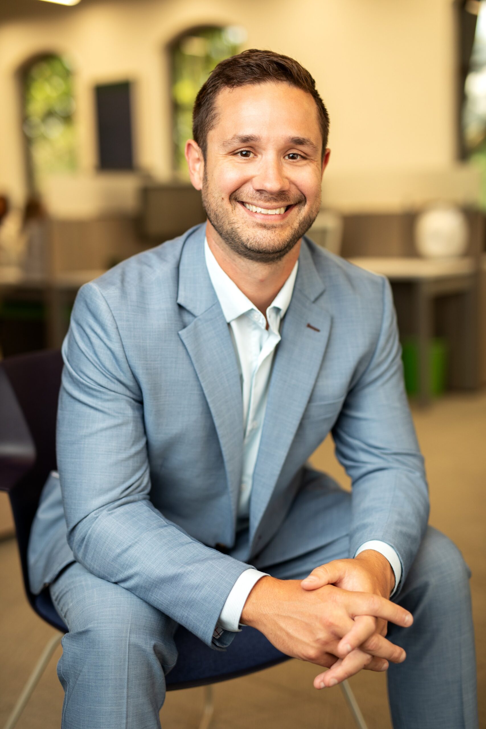 A man in a light blue suit is sitting and smiling in a modern office setting with blurred background.