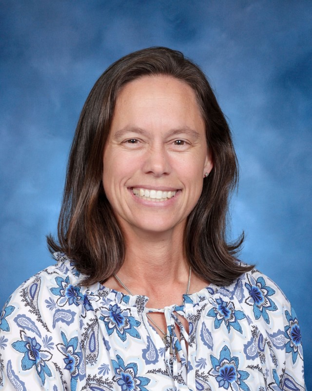 Woman with shoulder-length brown hair wearing a floral blouse, smiling in front of a blue studio background.