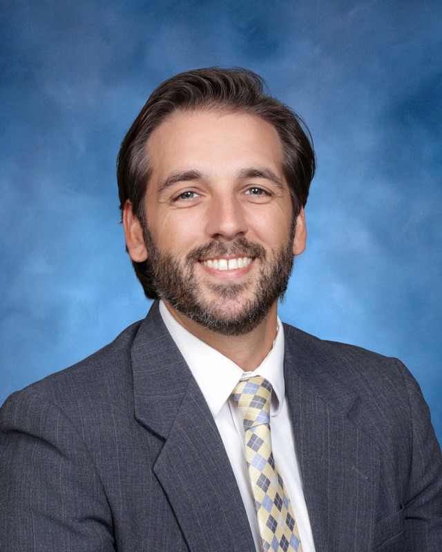A man in a gray suit, white shirt, and plaid tie smiles at the camera against a blue studio background.