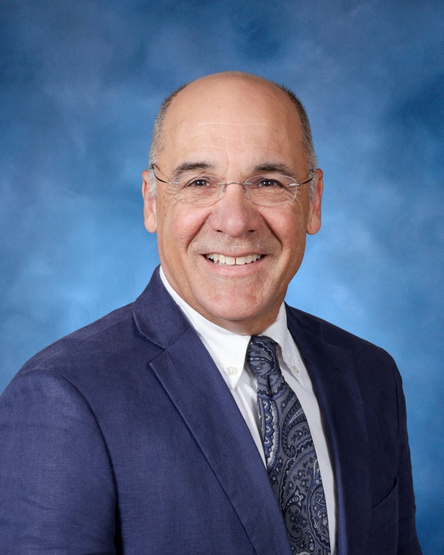 A middle-aged man wearing glasses, a blue suit, white shirt, and patterned tie, smiling in front of a blue studio background.