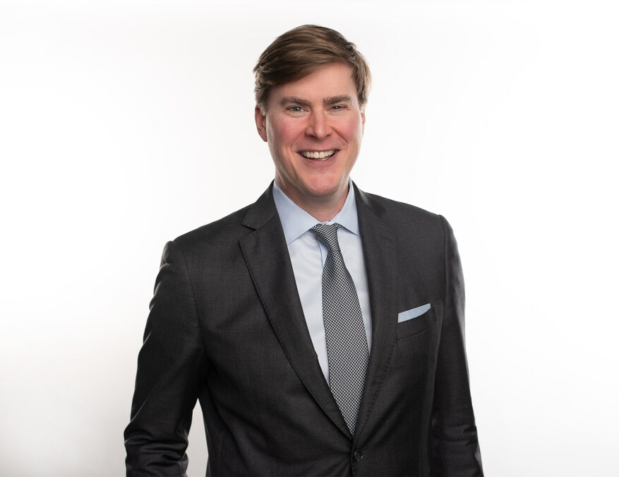 A man in a dark suit, light blue shirt, and patterned tie stands smiling against a plain white background.