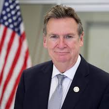 A man in a suit and tie stands in front of a blurred American flag, looking at the camera and smiling slightly.