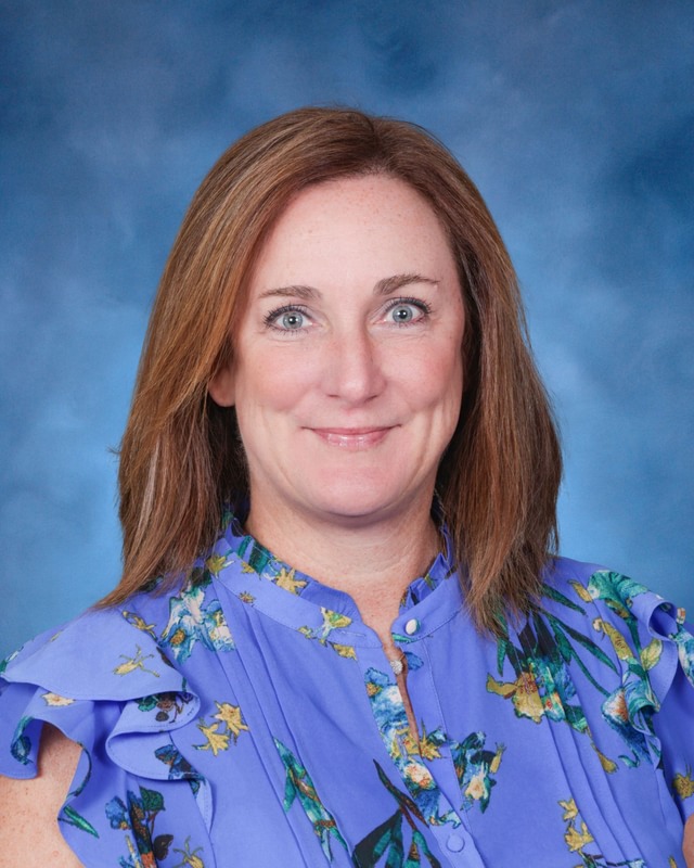 A woman with straight brown hair wearing a blue floral blouse poses in front of a blue studio background.
