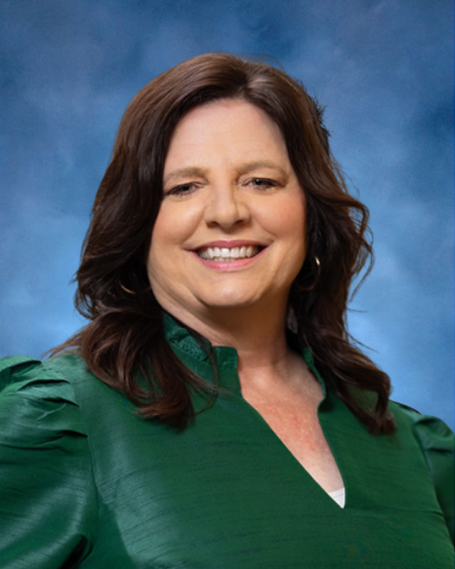 A woman with medium-length brown hair in a green blouse smiles, representing alumni relations, against a blue studio background.