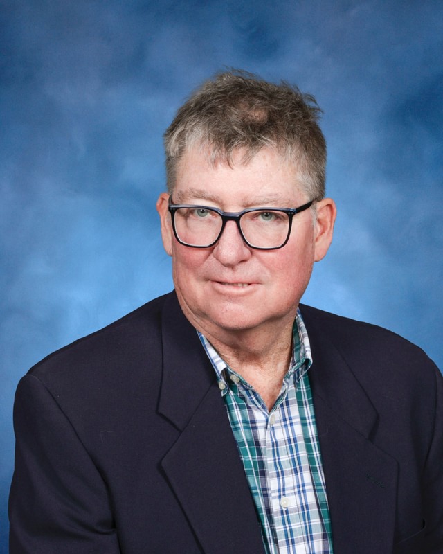 A man with short gray hair and glasses wearing a dark blazer and plaid shirt poses against a blue background.