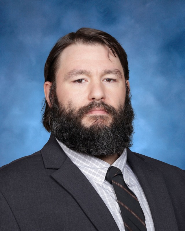 A man with a full beard and dark hair wearing a suit, striped tie, and checked shirt poses in front of a blue background.