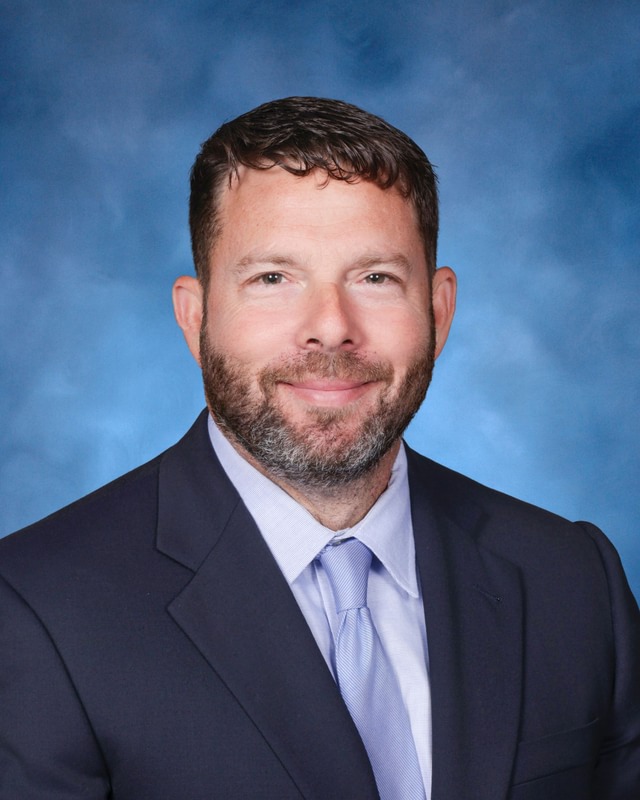 A man with short brown hair and a beard wearing a dark suit, light blue shirt, and tie poses in front of a blue studio background.