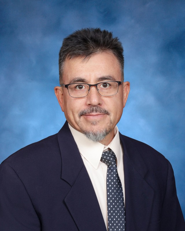 A man in a suit and tie with glasses and short dark hair poses for a formal portrait against a blue background.