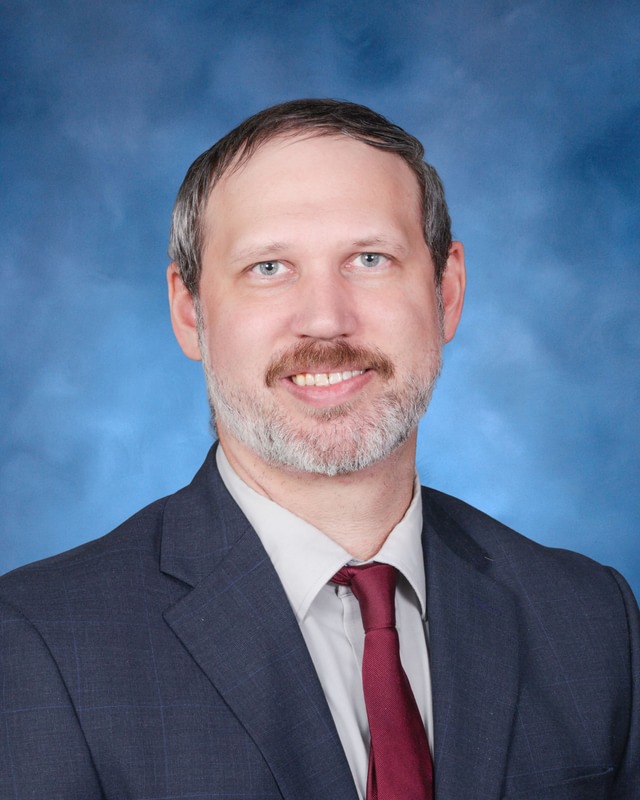 A man in a suit and tie poses for a formal portrait against a blue studio background.