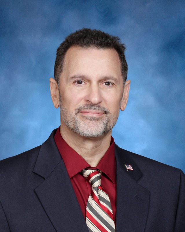 A man with short brown hair, trimmed beard, wearing a dark suit, red shirt, and striped tie poses against a blue studio backdrop.