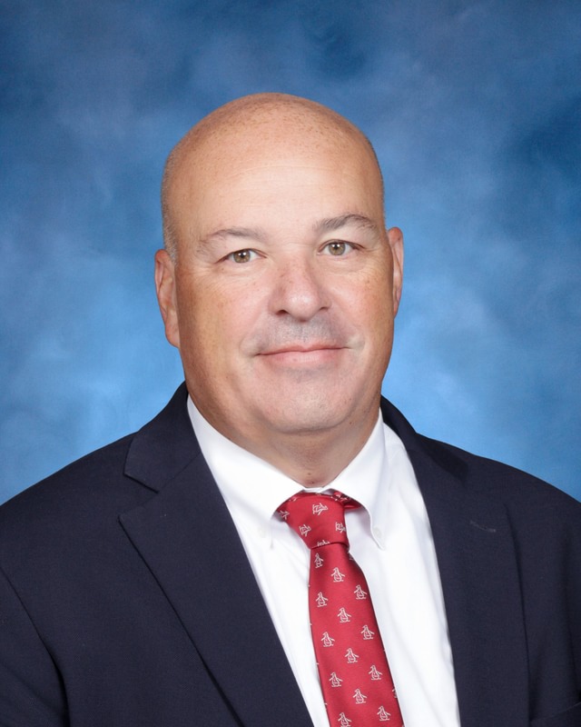 A man in a suit and red tie poses for a formal portrait against a blue background.