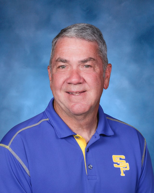 An older man with gray hair wears a blue and yellow collared shirt with "SP" letters, posed in front of a blue background.