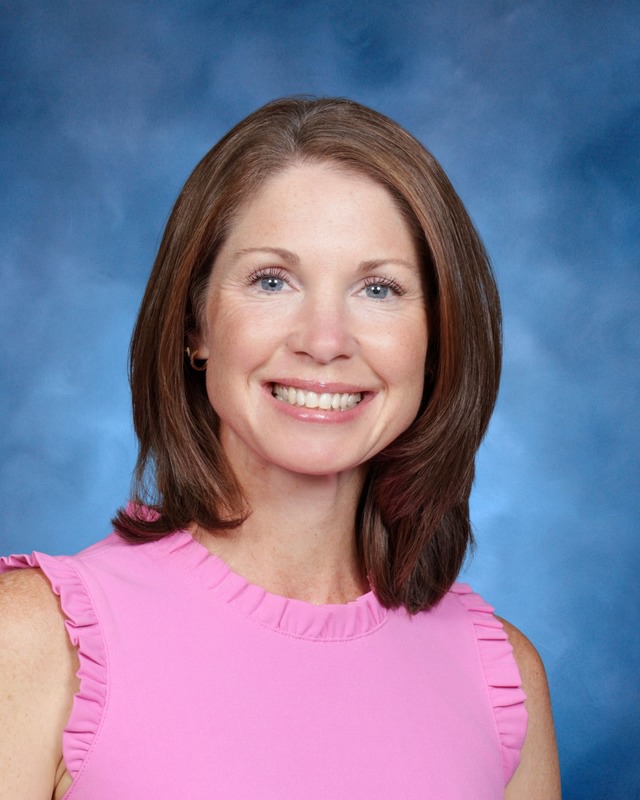 A woman with straight brown hair and a pink sleeveless top smiles in front of a blue studio background.