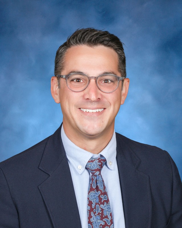 A man wearing glasses, a dark suit, a light blue shirt, and a patterned tie poses in front of a blue studio background.
