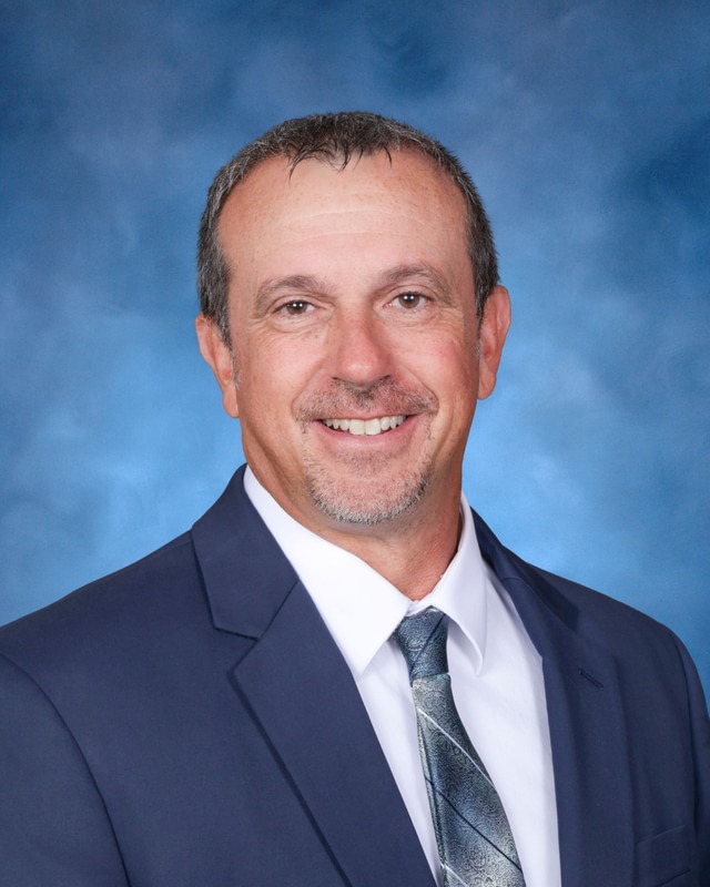 A middle-aged man in a navy suit, white shirt, and patterned tie smiles in front of a blue background.