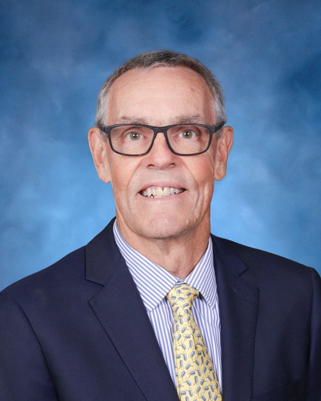A man with short gray hair and glasses wearing a navy suit, striped shirt, and yellow patterned tie against a blue background.