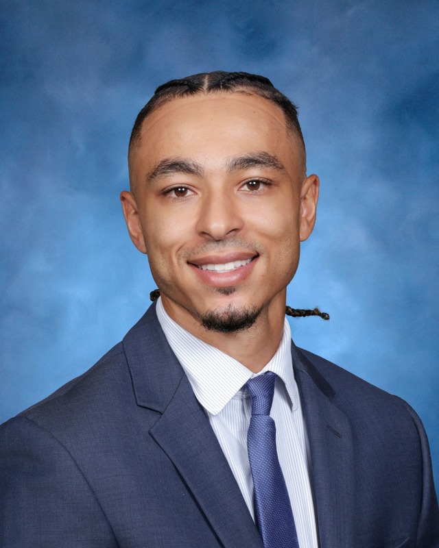 Man in a dark suit and tie with a light shirt, smiling, posed in front of a blue background.