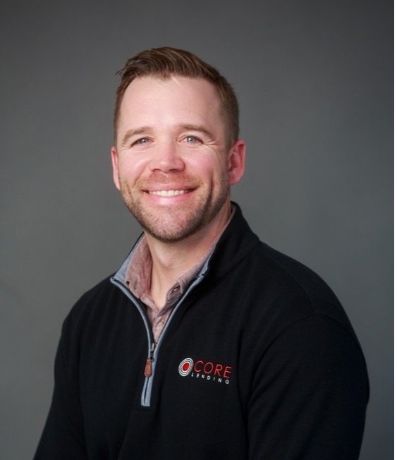 A man with short hair and a beard is smiling, wearing a black Core Lending pullover, in front of a gray background.