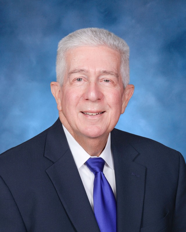 An older man with short gray hair in a dark suit, white shirt, and blue tie poses in front of a blue backdrop.