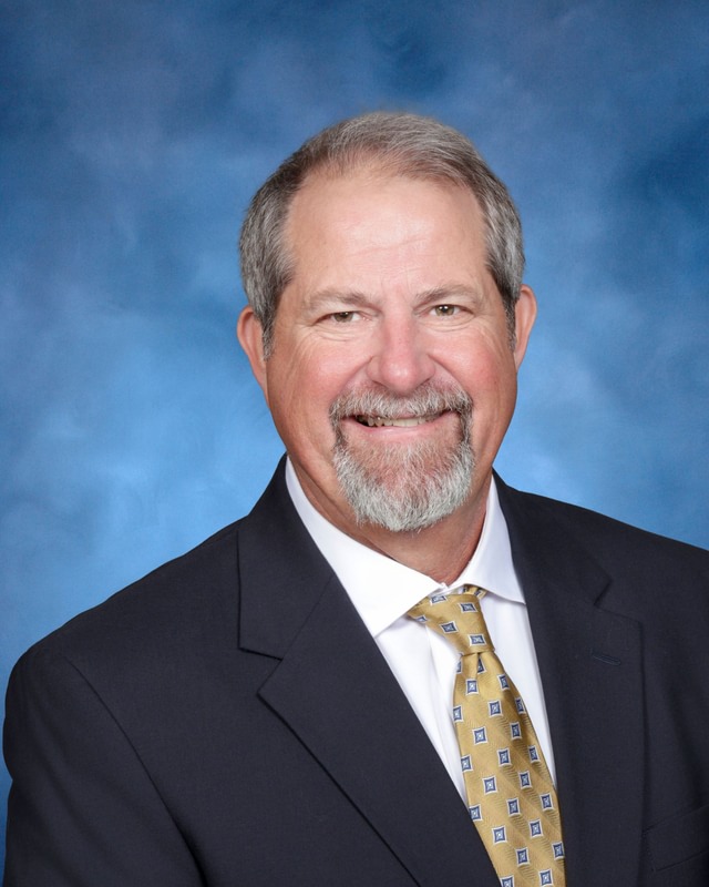 Man with gray hair and beard wearing a dark suit, white shirt, and yellow patterned tie, posing in front of a blue background.