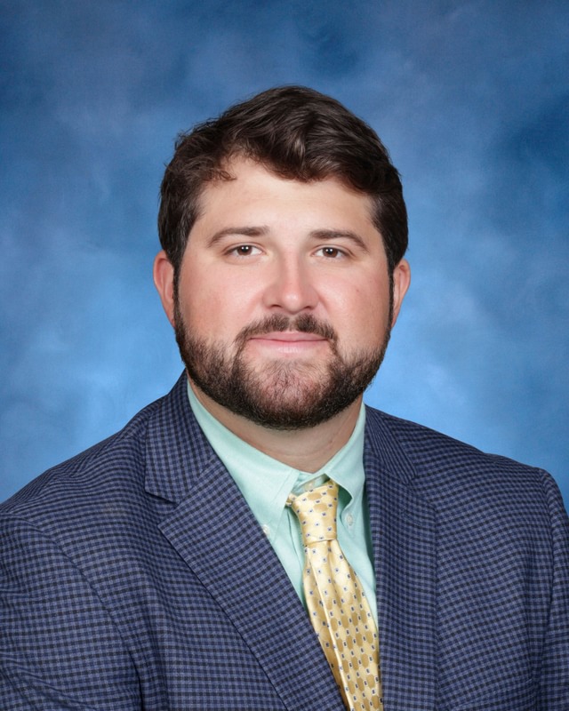 A man with short brown hair and a beard wearing a checked suit jacket, mint green shirt, and yellow tie poses in front of a blue background.