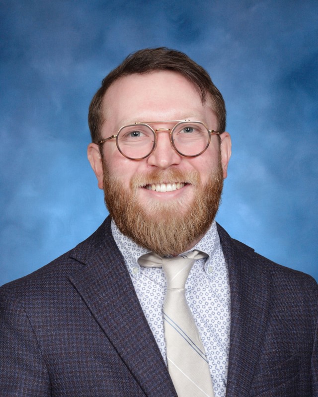 A man with glasses and a beard wears a blue suit, patterned shirt, and light tie, smiling in front of a blue background.
