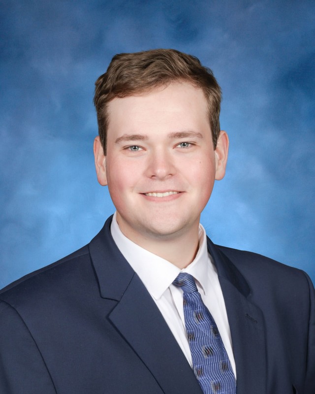 A young man in a navy suit, white shirt, and patterned tie poses for a formal portrait against a blue studio background.
