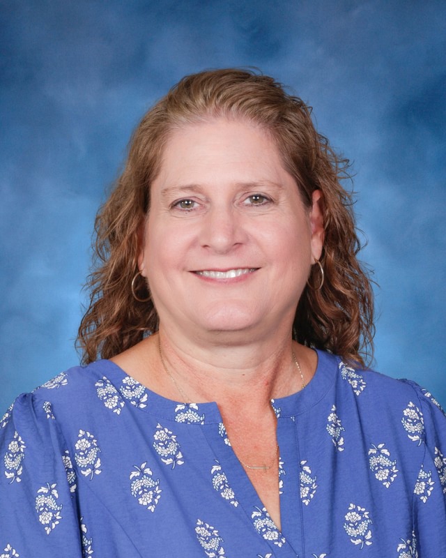 A woman with wavy light brown hair wearing a blue patterned blouse, smiling in front of a blue studio background.
