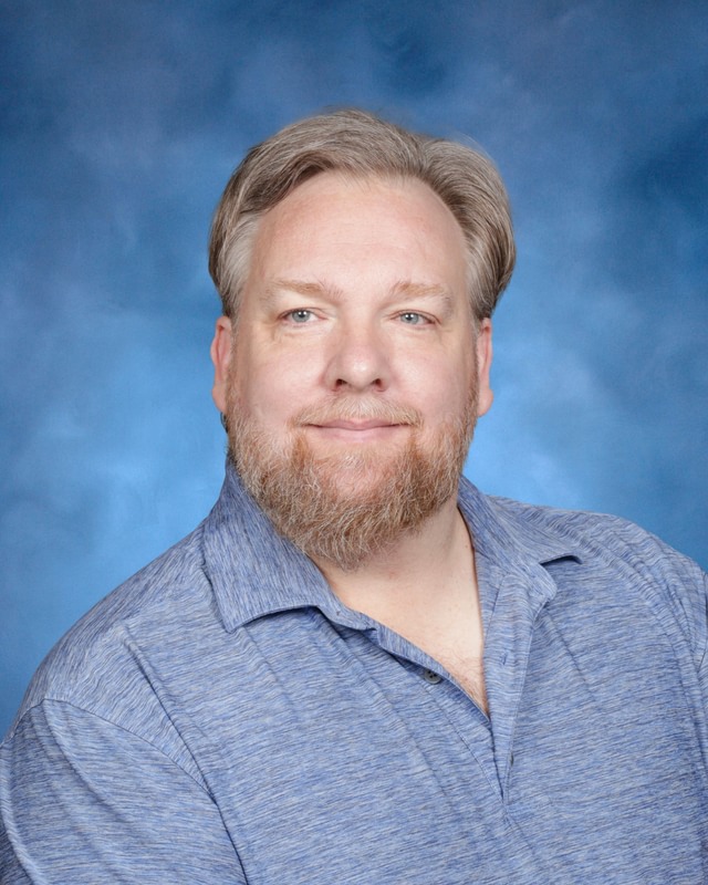 A middle-aged man with light hair and a beard wearing a blue collared shirt poses in front of a blue studio background.