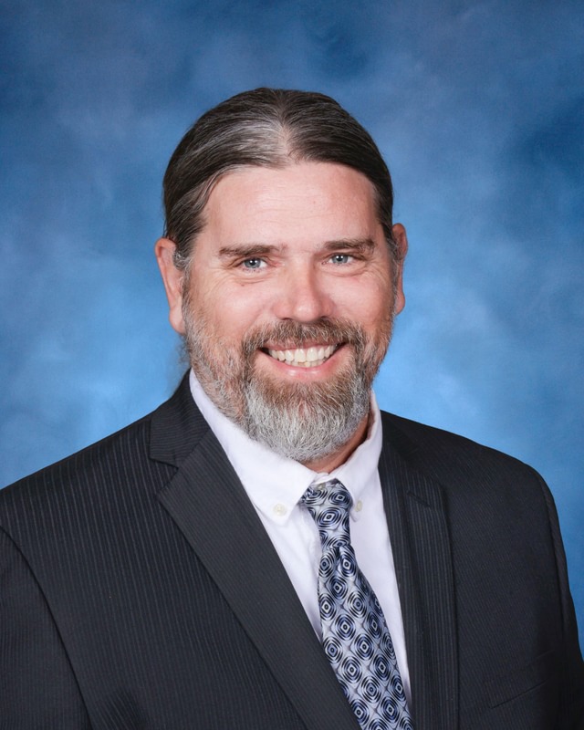 A man with gray hair and beard, wearing a dark suit, white shirt, and patterned tie, smiles in front of a blue background.