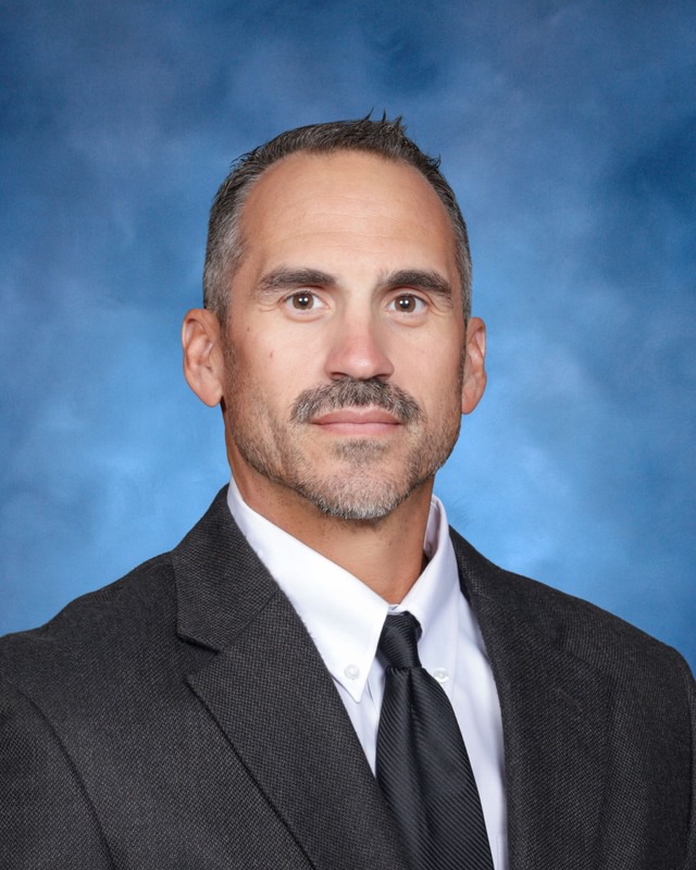 A man with short dark hair and a trimmed beard wearing a dark suit, white shirt, and black tie poses in front of a blue background.