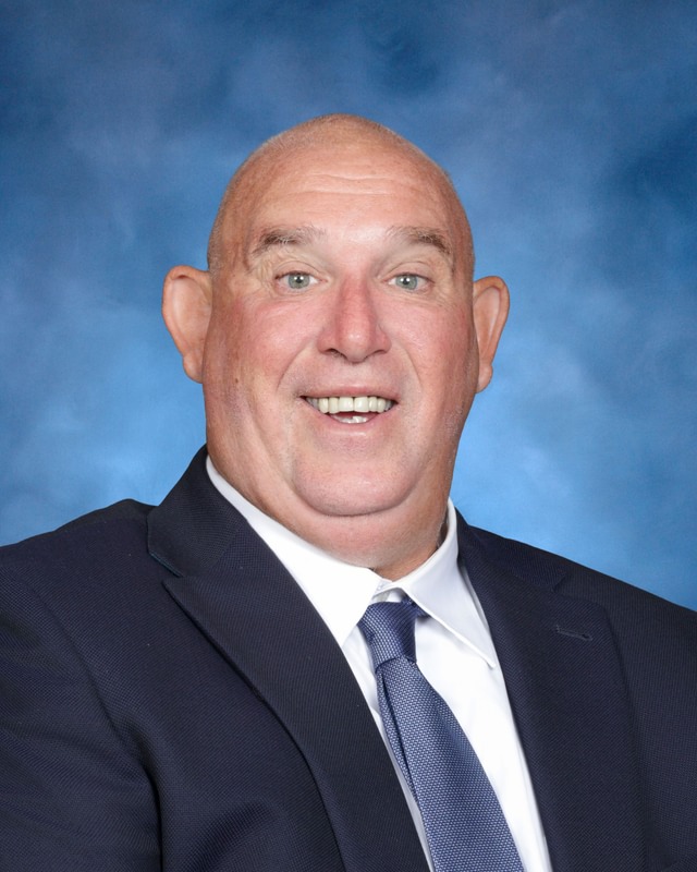 A bald man in a dark suit, white shirt, and blue tie smiles in front of a blue studio background.