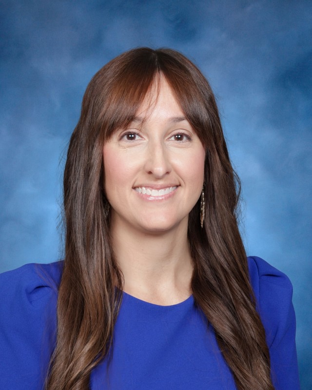 A woman with long brown hair wearing a blue top smiles at the camera in front of a blue background.
