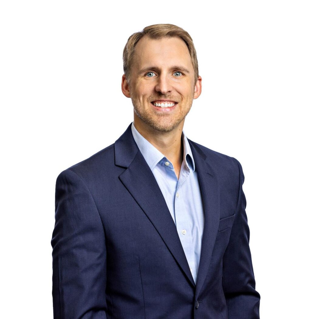 A man in a navy blue suit and light blue shirt is smiling at the camera against a plain white background.