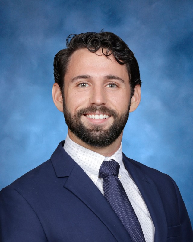 A man with dark hair and a beard wearing a navy suit, white shirt, and tie, smiling in front of a blue background.