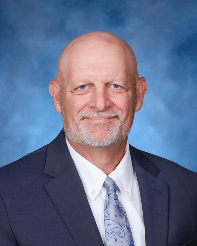 Bald man with a gray beard wearing a navy suit, white shirt, and patterned tie, posing in front of a blue backdrop.