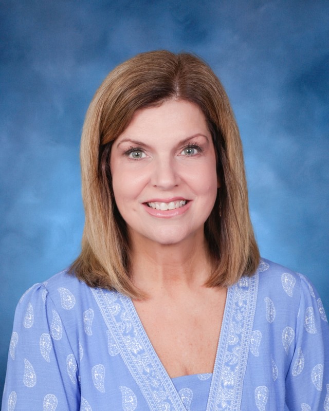 A woman with shoulder-length light brown hair smiles at the camera, wearing a light blue patterned blouse, with a blue background.