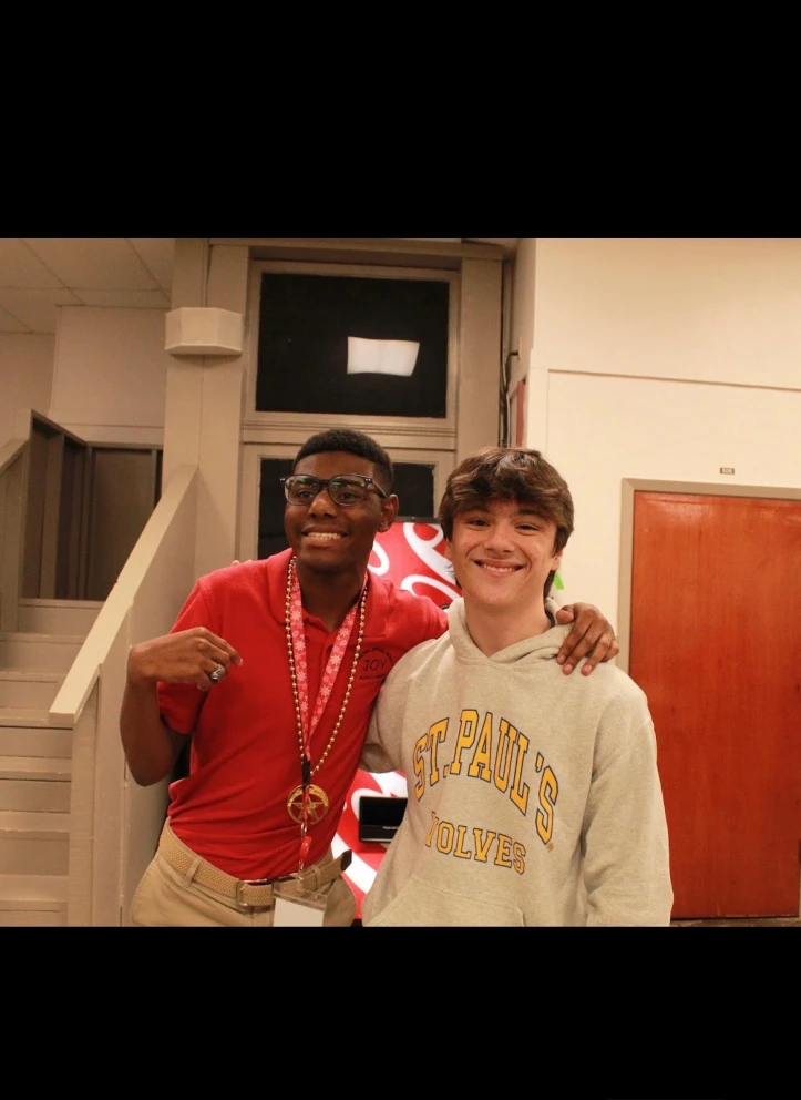 Two young men stand indoors, one in a red shirt with a medal, the other in a gray Saint Paul's hoodie, smiling after Sophomore Service Day.