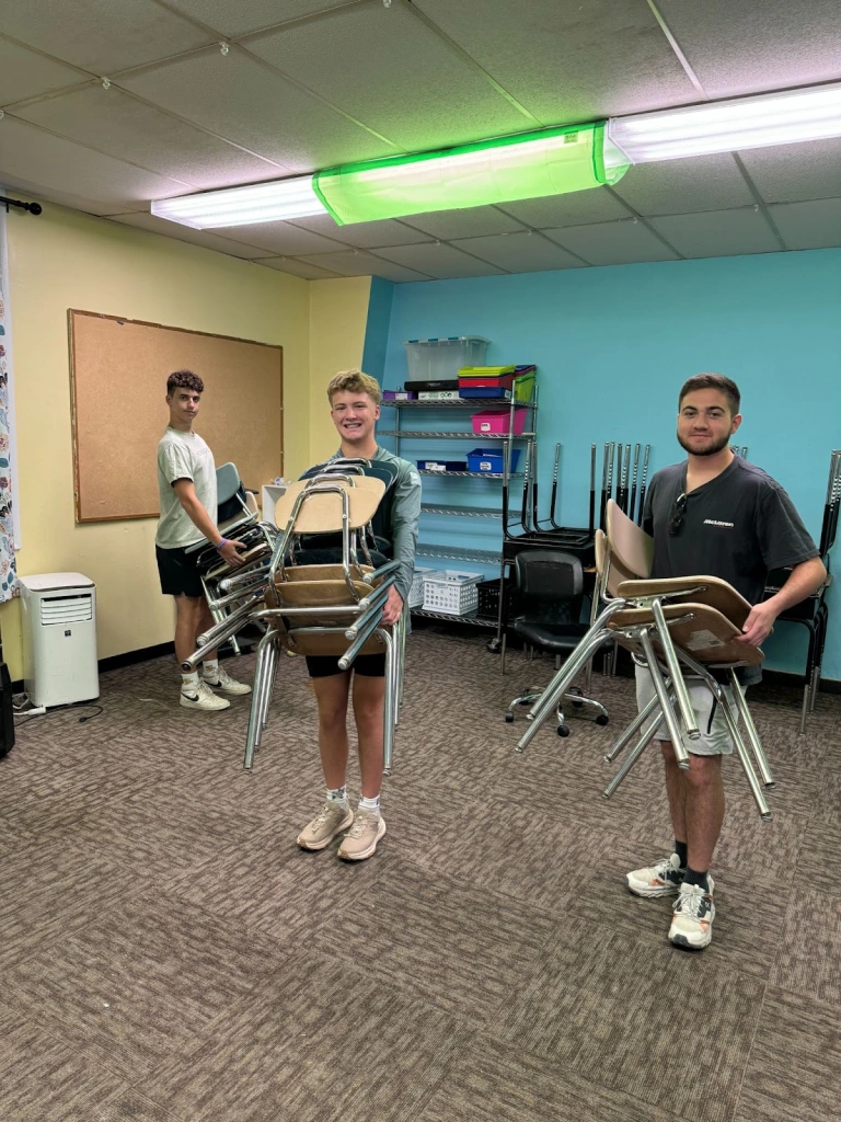 Three St. Paul's students carry stacks of chairs in a classroom with blue walls, shelves, and a bulletin board.