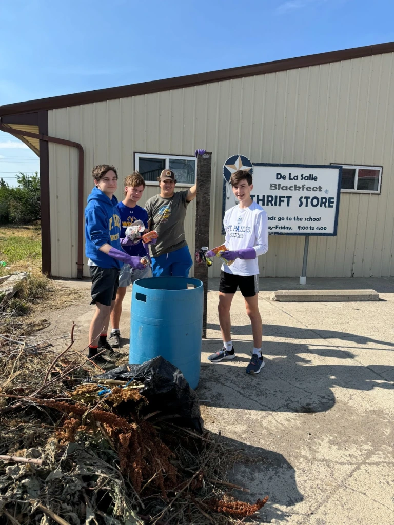 Four St. Paul's students wearing gloves clear debris into a blue bin outside De La Salle Blackfeet Thrift Store, embracing Blackfeet culture.