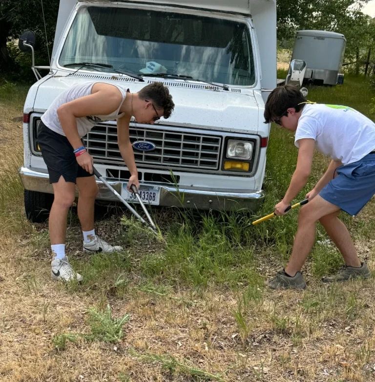 St. Paul's students use loppers to cut tall grass, embracing cultural immersion in front of a white Ford van on a grassy area.
