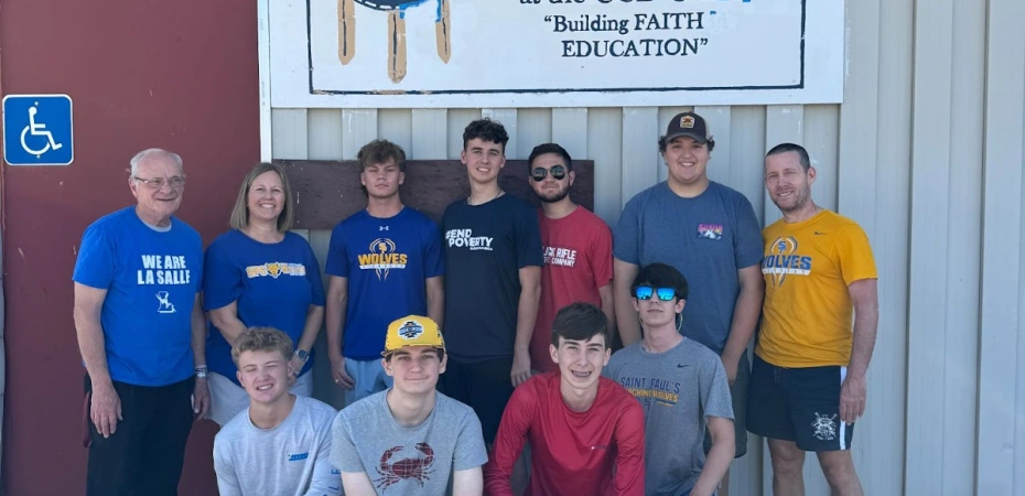 A group of ten St. Paul's students poses in front of a building labeled "Building FAITH EDUCATION." Some display pride in Blackfeet culture.