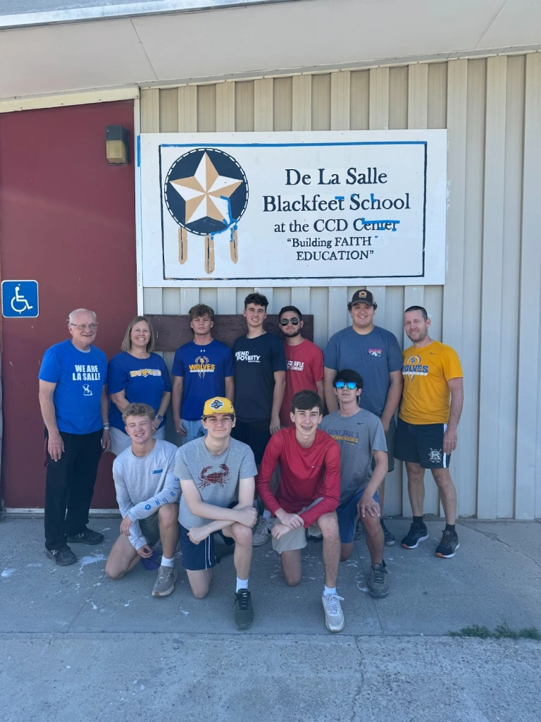 A group of eleven St. Paul's students pose in front of a De La Salle Blackfeet School sign, embracing Blackfeet culture on a sunny day.