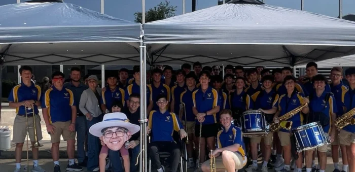 A large group in blue and yellow uniforms stand under canopies outdoors, some holding instruments, showing Phoenix spirit as they Rise Like Him.