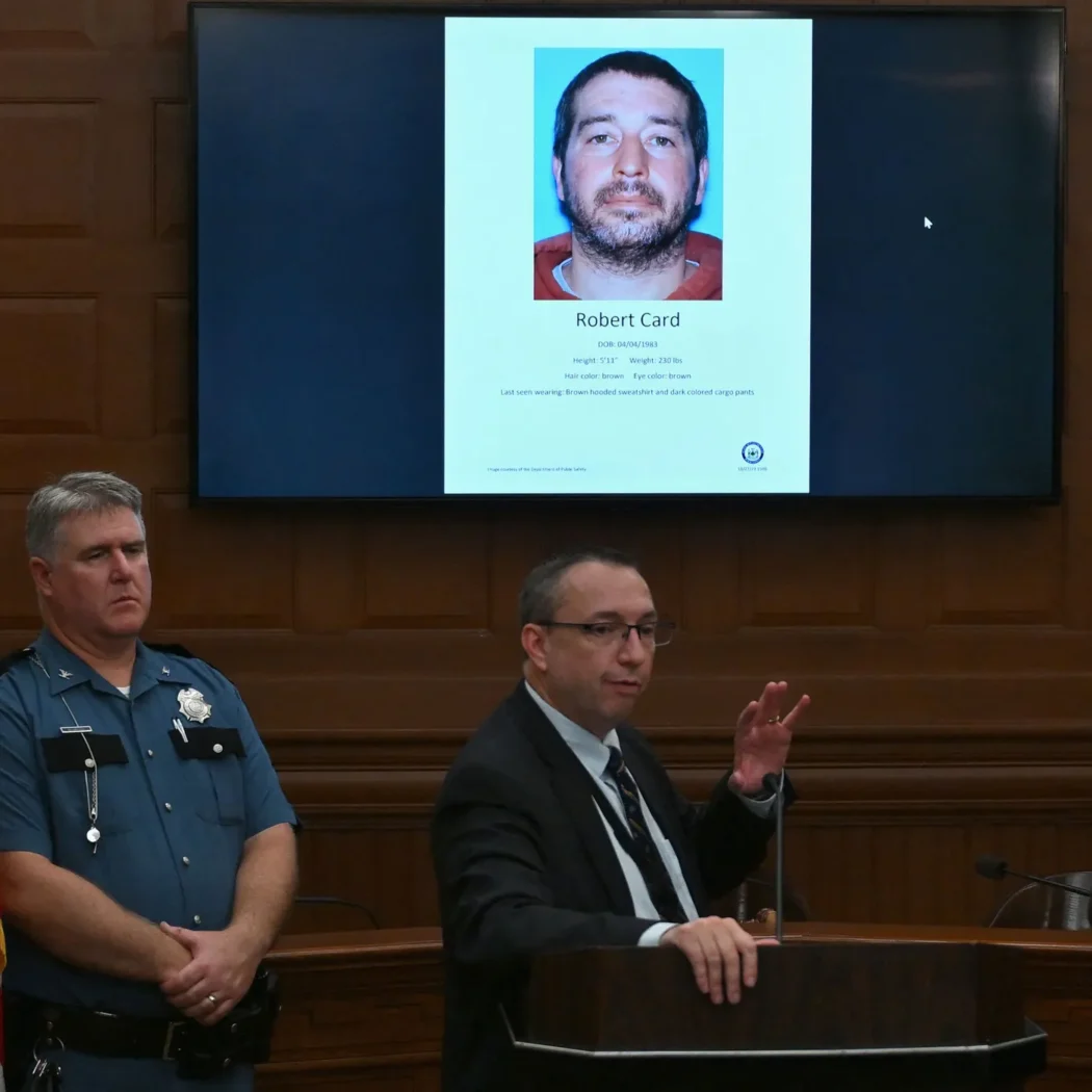 A man speaks at a podium in a courtroom; behind him, a screen displays a photo and information about Robert Card. A police officer stands nearby.