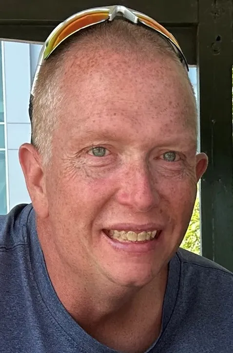 A man with short, light hair and freckles is smiling, wearing a blue shirt and sunglasses on his head, seated outdoors.